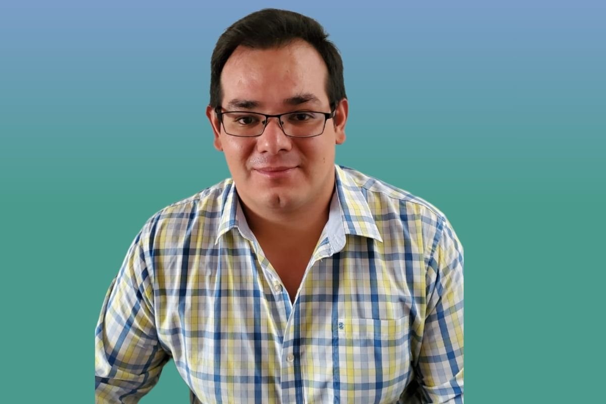 Hombre con lente de color negro y una camisa de cuadros de color amarilla con azul en un fondo de color azul con verde